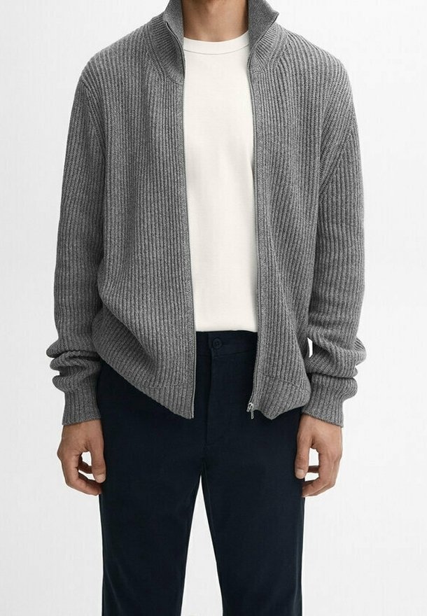 Cardigan grigio a zip con coste e colletto alto, indossato sopra una maglietta bianca, abbinato a pantaloni scuri. Texture in maglia, design aderente.