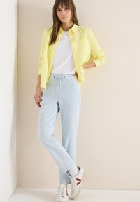 Femme debout contre un mur beige, vêtue d'une chemise jaune boutonnée, d'un t-shirt blanc, d'un pantalon bleu clair à cordon, de chaussettes blanches et de baskets blanches avec des accents rouges.