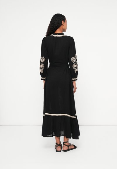 Fekete maxiruha hosszú ujjakkal, szőtt virágmintákkal fehér és krém színben, öv a deréknál és réteges alj, fekete szandálokkal párosítva.