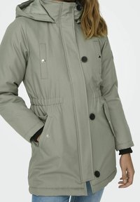 Ljusgrå, vadderad parka med huva, utrustad med markerad midja, svarta knappar och kontrasterande mörka muddar. Har framfickor.