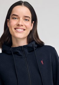 Sudadera con cremallera en color navy, hecha de algodón suave, con cordones y un pequeño logo rojo en el pecho. Presenta un cuello alto y un ajuste ceñido.