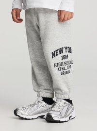 Pantaloni in pile grigi con testo stampato nero: "NEW YORK 1984 HIGH SCHOOL ATHL. DEPT. ORIGINAL." Polsini elasticizzati. Paio di scarpe sportive argento.
