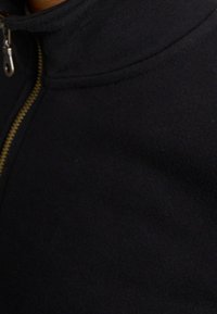 Detailní záběr horní části černé zipové bundy, zobrazující táhlo zipu a texturované detaily látky.