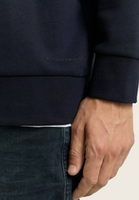 Sudadera azul marino hecha de una tela suave, con puños acanalados, cuello redondo y un logotipo en relieve en el dobladillo. Combinada con jeans oscuros.