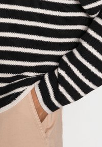 Černobílý pruhovaný svetr s texturovaným pletením, který má široké pruhy a uvolněný střih. Světlé kalhoty jsou viditelné v oblasti kapes.