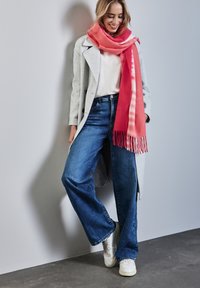 Manteau en laine gris, pull blanc, écharpe dégradé rose, jean bleu à pattes d'éléphant et baskets blanches. Textures lisses avec des franges sur l'écharpe.