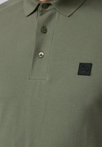 Oliwkowa koszulka polo wykonana z teksturowanej tkaniny, z kołnierzykiem, trzema guzikami i czarną prostokątną łatą z logo na piersi.