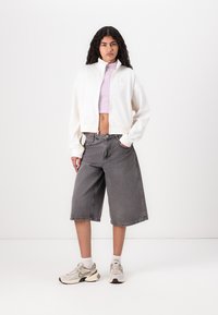 Hvid cropped jakke med høj krave, parret med grå denim shorts til knæene. Modellen bærer lys grå sneakers og en lilla top.
