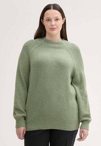 Grüner gerippter Pullover mit langen Ärmeln und rundem Halsausschnitt, mit lockerer Passform und strukturiertem Stoff. Getragen über schwarzen Hosen.