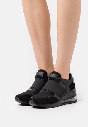 Schwarze Sportschuhe mit einer Mischung aus glatten und strukturierten Materialien, ausgestattet mit breiten Klettverschlüssen, einer geschwungenen Sohle und einem transparenten Fersenbereich.