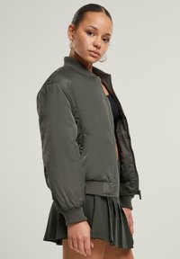 Dunkelgrüne Bomberjacke mit geripptem Bund und Bündchen, ausgestattet mit einem Frontreißverschluss und Seiten Taschen, getragen über einem schwarzen Oberteil und einem Rock.