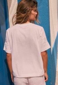 T-shirt en coton blanc à manches courtes, avec un détail de broderie florale sur la manche gauche. Design simple et décontracté, texture douce.