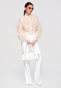Femme portant un chemisier beige transparent avec des appliqués floraux, un pantalon blanc, des lunettes de soleil roses et tenant un sac à main blanc sur un fond uni.