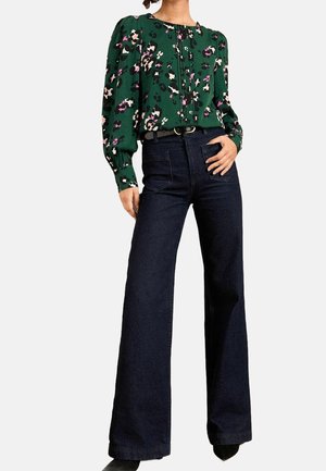 Femme portant un chemisier vert à fleurs avec des manches longues bouffantes et un pantalon large sombre taille haute, debout avec une main sur la hanche.