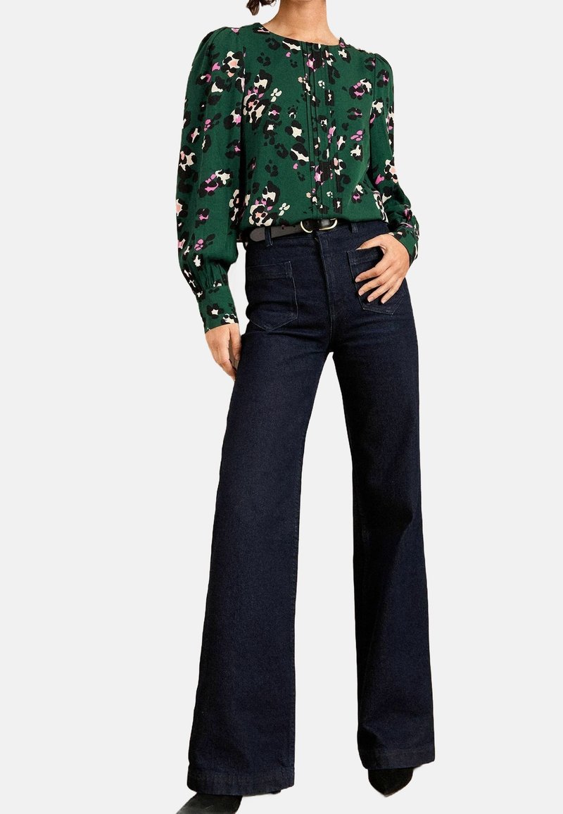Femme portant un chemisier vert à fleurs avec des manches longues bouffantes et un pantalon large sombre taille haute, debout avec une main sur la hanche.