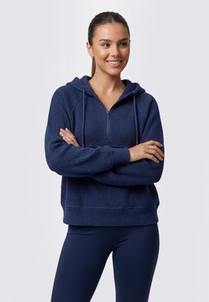 Junge Frau steht mit verschränkten Armen, lächelnd, trägt einen marineblauen, gerippten Zip-Hoodie und marineblaue Leggings vor einem einfarbigen hellen Hintergrund.