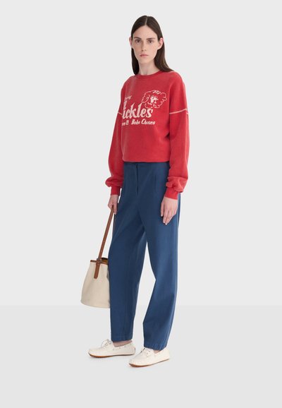 Femme aux longs cheveux noirs portant un sweat-shirt rouge à motifs, un pantalon taille haute bleu, des chaussures blanches, et tenant un sac à main beige avec des anses marron.