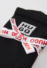 Calze nere a coste con un'etichetta bianca con il logo "HUGO" e un nastro bianco attorno, stampato con "HUGO" in rosso.