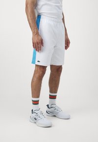 Man som bär vita och ljusblå träningsshorts, vita höga strumpor med gröna och röda ränder, samt vita Lacoste-sneakers på en enfärgad bakgrund.