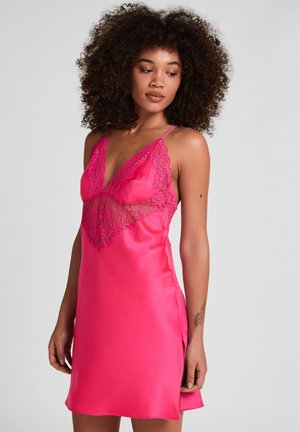 Vrouw met krullend haar die een felroze satijnen nachtjapon met kantdetail op het lijfje draagt, kijkend naar beneden links van haar.
