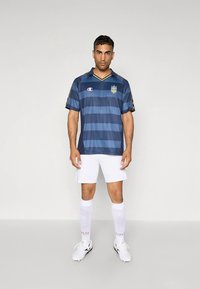 Tamsiai mėlynas futbolo marškinėlis su horizontaliomis juostelėmis, trumpomis rankovėmis ir apykakle. Dėvimas su baltais šortais ir baltomis kelių ilgio kojinėmis, ant kurių matomi logotipai.