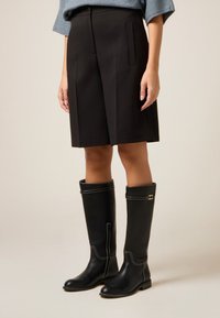 Svarta knähöga läderstövlar med kontrasterande vit söm och en liten guldfärgad detalj, kombinerade med skräddarsydda svarta shorts.