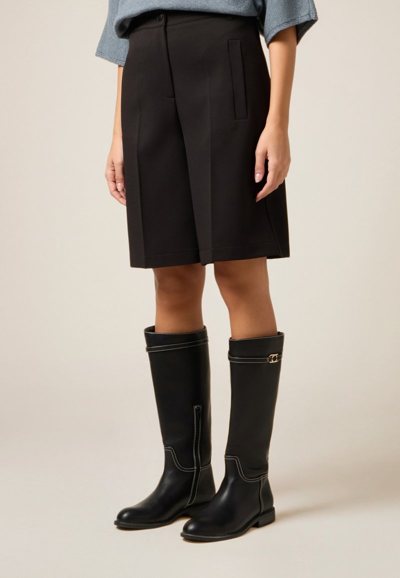 Svarta knähöga läderstövlar med kontrasterande vit söm och en liten guldfärgad detalj, kombinerade med skräddarsydda svarta shorts.