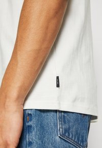 Weißes Baumwoll-T-Shirt mit kurzen Ärmeln, seitlichem Etikett, getragen mit blauen Jeans aus Denim mit orangefarbenen Nähten.