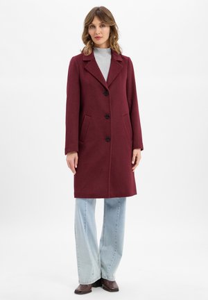 Cappotto di lana bordeaux con colletto a punte, tre bottoni, due tasche laterali e maniche lunghe, abbinato a jeans a gamba larga di un blu chiaro.