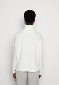 Weißer, gerippter Rollkragenpullover mit entspanntem Schnitt, fällt mit überschnittenen Schultern und langen Ärmeln. Weiche Textur; minimalistische Gestaltung.