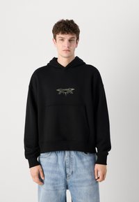 Schwarzer Hoodie mit einem Kängurutasche, der ein dekoratives Logo in der Mitte aufweist. Der Stoff wirkt weich und hat einen lockeren Schnitt sowie gerippte Bündchen.