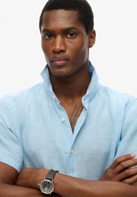 Hombre con los brazos cruzados, vestido con camisa azul claro y reloj de pulsera negro, mostrando un tatuaje en la parte superior del pecho.