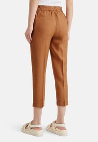 Hnědé lněné kalhoty s elastickým pasem, zkrácenou délkou, ohrnutými manžetami a texturovaným povrchem. Nosič má na sobě bílé sandály.