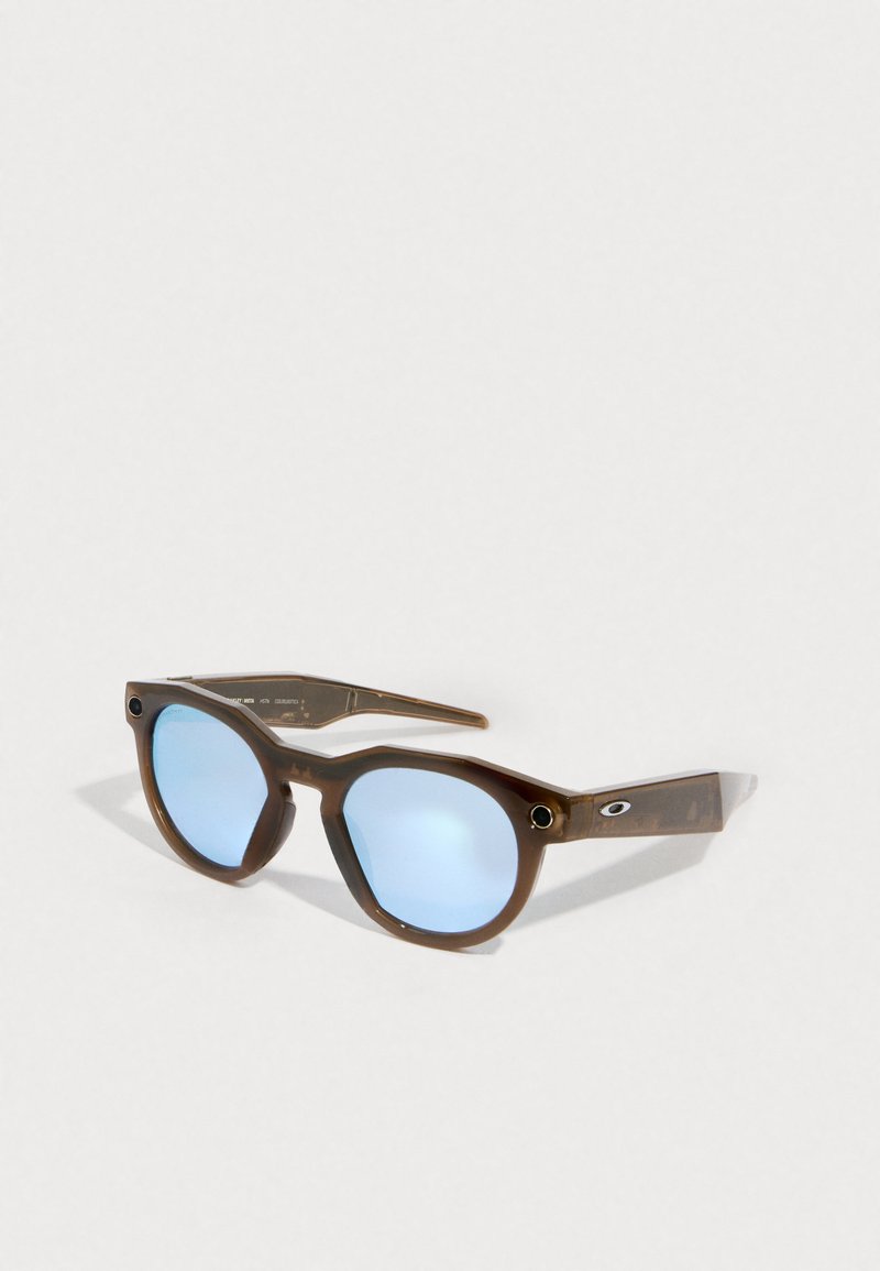 Brown plastic sunglasses with round blue lenses, featuring a unique design, patterned frames, and small silver hardware accents on the sides.