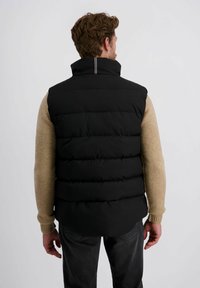 Schwarzes gepolstertes Weste mit hohem Kragen, versehen mit einem weißen Streifendetail auf der Rückseite, getragen über einem beigen Pullover, sichtbar mit grauen Jeans.
