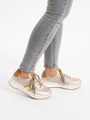 Legs wearing light gray skinny jeans and beige lace-up platform sneakers with yellow and brown accents, standing on a white surface.