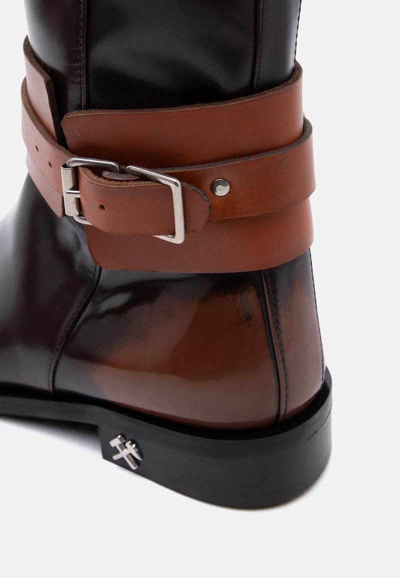 Black and brown leather boot with a thick brown strap and silver buckle around the ankle, featuring a small metal emblem on the sole.