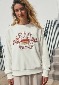Biała bluza z napisem "Sweater Weather" i ilustracją postaci. Długie rękawy, ściągacze przy mankietach i u dołu. Gładka tekstura.