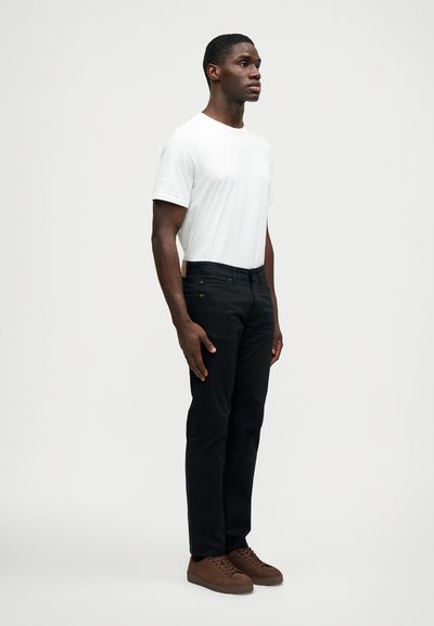 Άνδρας που στέκεται φορώντας ένα λευκό T-shirt, μαύρα τζιν και καφέ αθλητικά παπούτσια μπροστά σε έναν απλό λευκό φόντο.