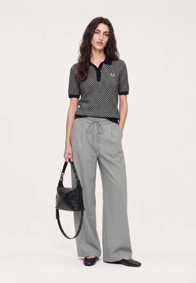 Femme aux longs cheveux foncés debout, portant un polo à motif zigzag noir et gris, un pantalon gris à jambes larges, des ballerines noires, et tenant un sac à main noir.