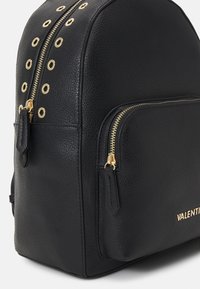 Svart läderryggsäck med gyllene detaljer, med en texturerad yta, dubbla dragkedjor och dekorativa gyllene öglor på sidan.