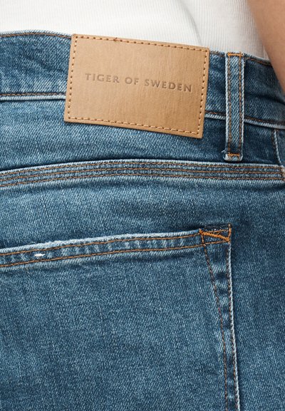 Κοντινή λήψη της πίσω τσέπης από μπλε τζιν με ετικέτα από ταμπά δέρμα με το λογότυπο "Tiger of Sweden" ραμμένο πάνω από την τσέπη.