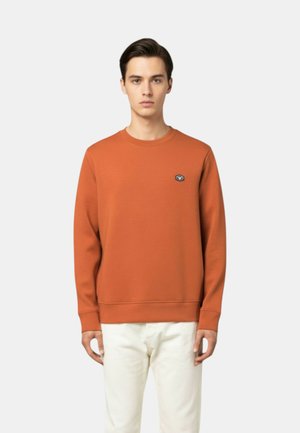 Giovane uomo con capelli corti e scuri che indossa una felpa girocollo color ruggine senza decorazioni e pantaloni bianco sporco, in piedi davanti a uno sfondo bianco.