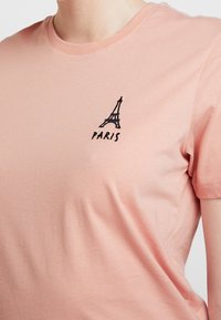 Růžové bavlněné tričko s malou černou vyšívanou Eiffelovkou a slovem "PARIS" pod designem. Krátké rukávy a kulatý výstřih.