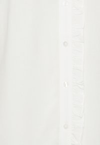 Camicia bianca con bottoni frontali trasparenti e dettagli arricciati lungo il centro. Tessuto morbido.
