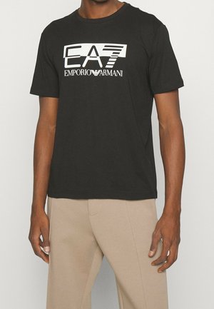 Mand iført sort EA7 Emporio Armani logo t-shirt og lyse beige bukser, stående mod en ensfarvet lys baggrund.