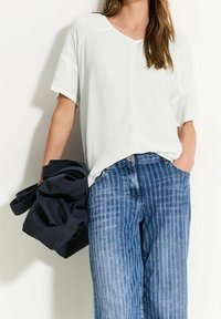 Vit kortärmad topp med V-ringning och texturerade axeldetaljer, kombinerad med blå randiga vida jeans och ett vikt plagg i mörk marinblå.