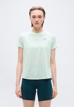 Jonge vrouw die een licht muntgroen kort mouw sportshirt en donker teal aansluitende shorts draagt, staand tegen een effen witte achtergrond.