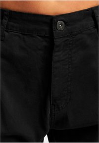 Zwarte katoenen broek met een soepele textuur, voorzien van een centrale knoopsluiting en zijzakken. Het stiksel is zichtbaar nabij de tailleband.