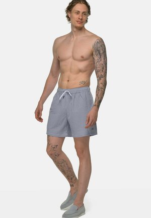 Oberkörperfreier Mann mit Tätowierungen am rechten Arm und linken Bein, trägt grau-weiß gestreifte Shorts und graue Slipper, steht vor weißem Hintergrund.